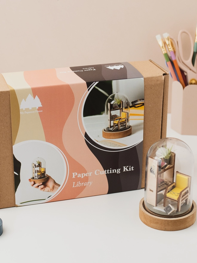 My Papercut Forest packaging box with the finished project in front of it. It's a mini library scene on a plastic dome.