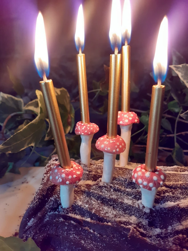 Five lit gold candles held in red-and-white ceramic mushroom candle holders on a chocolate log cake, styled for an enchanted woodland celebration.