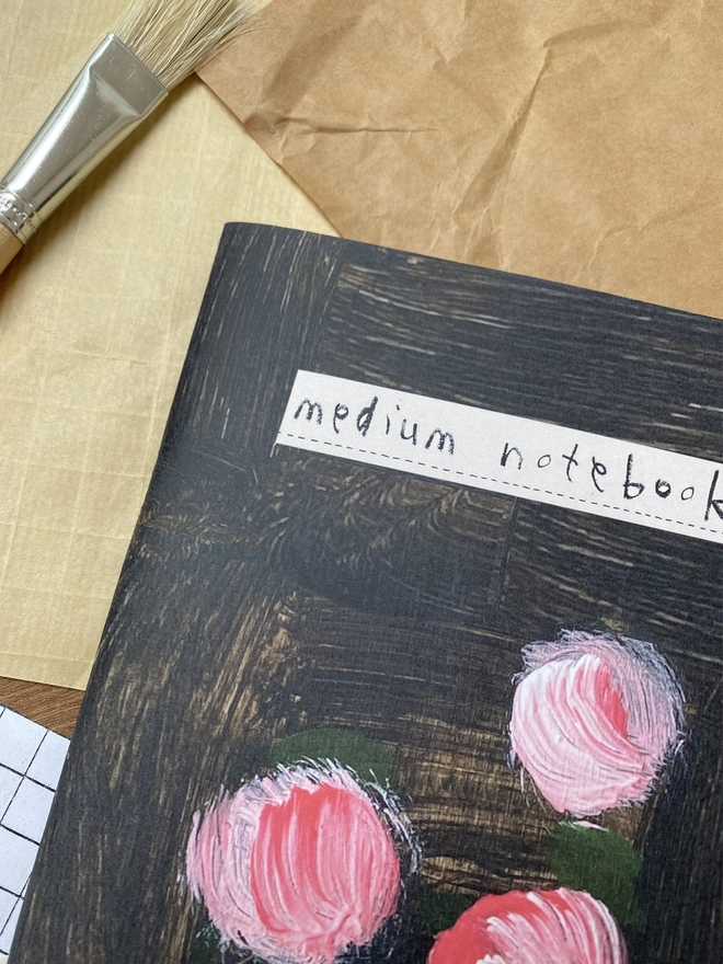 Notebook with dark cover and pink flowers on a desk with paintbrush.