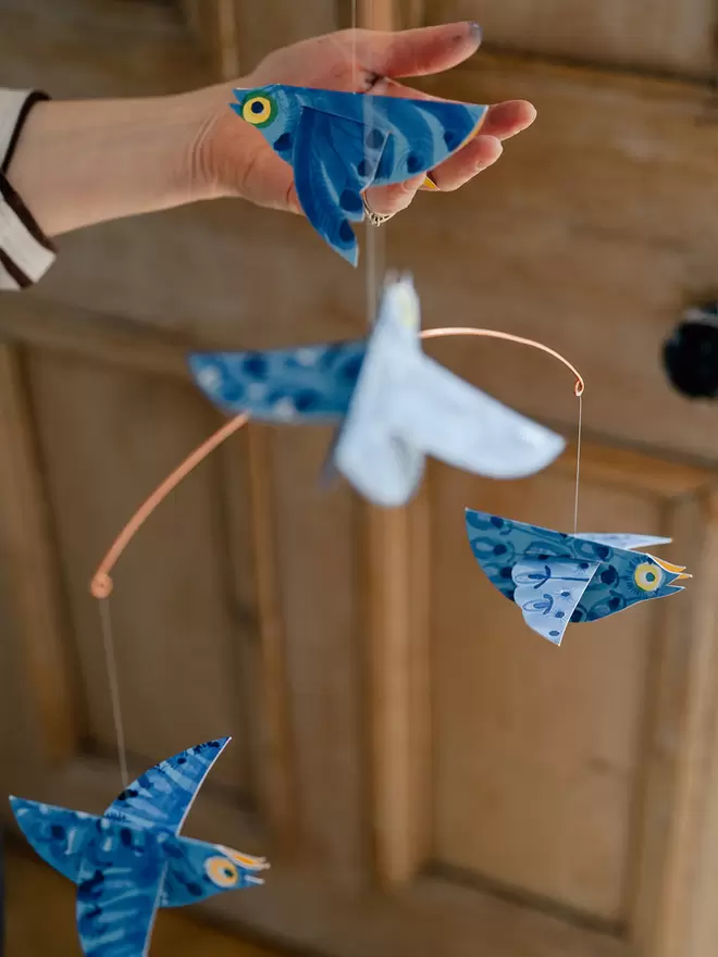 Bluebirds patterned paper birds mobile. Photo Zoe Barrie. hanging against a rustic wooden door. The artists' hand cups one bird.