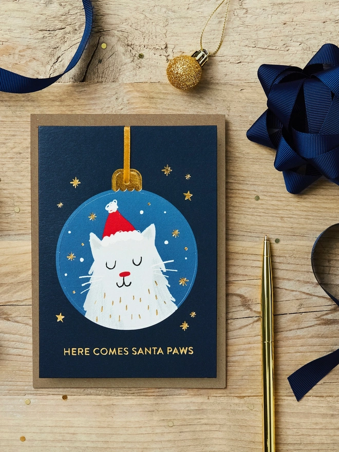 Christmas pop-out bauble card with a cat illustration on it sat on a wooden desk with blue and gold Christmas decorations and stationery.