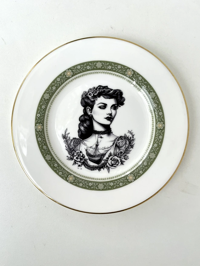 a beautifully ornate plate and in the centre is a vintage image of a beautiful tattooed london lady, she has ringlets and a choker and a tattooed ship on her chest
