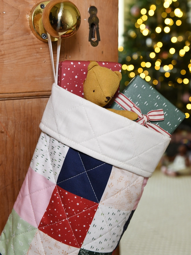 A patchwork stocking in festive colours with an ivory turnover cuff hangs on a wooden door. Small gifts and toys poke out of the top.