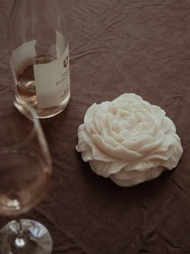 Large White Peony Candle. A top-down view of an ivory peony flower candle on a crumpled brown linen tablecloth, beside a glass and a bottle of rosé. The sculpted blooming petals add a romantic, refined touch to evening entertaining.