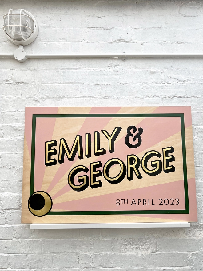 Modern wedding sign featuring the couple's names in gold leaf, outlined in black, and the wedding date, with a sunburst background and a border. Shown against a white brick wall, straight on. 