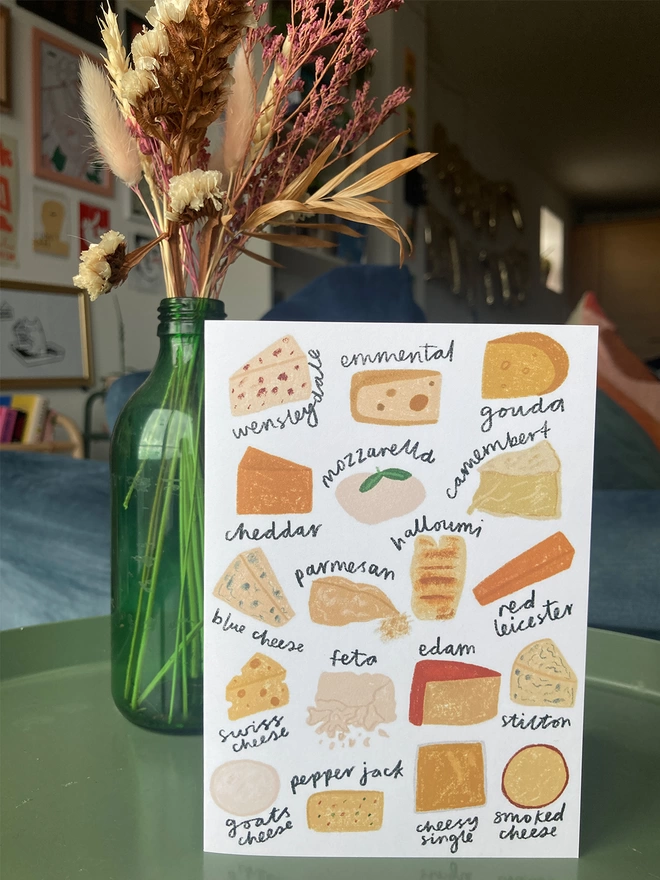 Greetings card illustrated with a variety of cheeses. Placed on a table with flowers in the background.