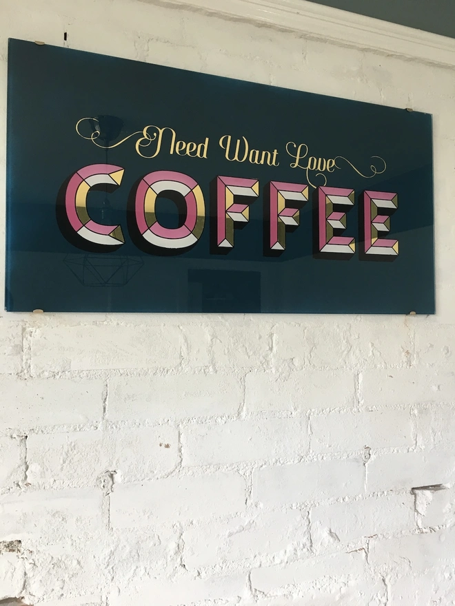 'Need Want Love Coffee' Gold Glass Sign, a gold glass sign hanging on a white brick wall. 