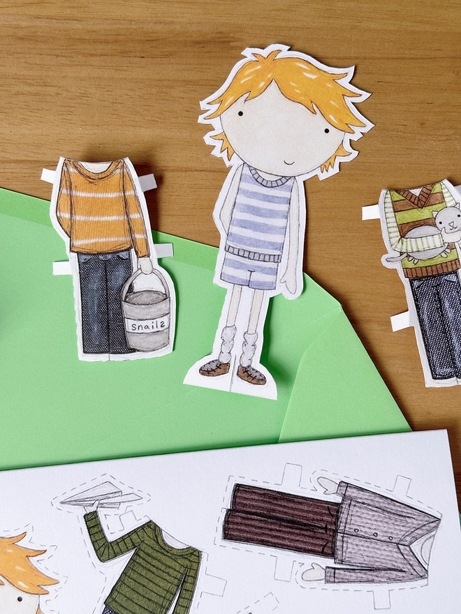 A paper doll greetings card with a paper doll and several outfits on the card lays on a green envelope on a wooden desk.