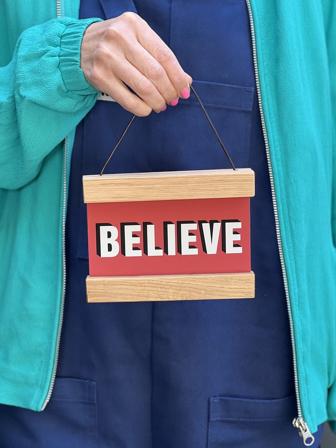 A print in red with the word BELIEVE framed in a oak hanger, with a waxed cotton cord for hanging being held by a hand with pink nail varnish 