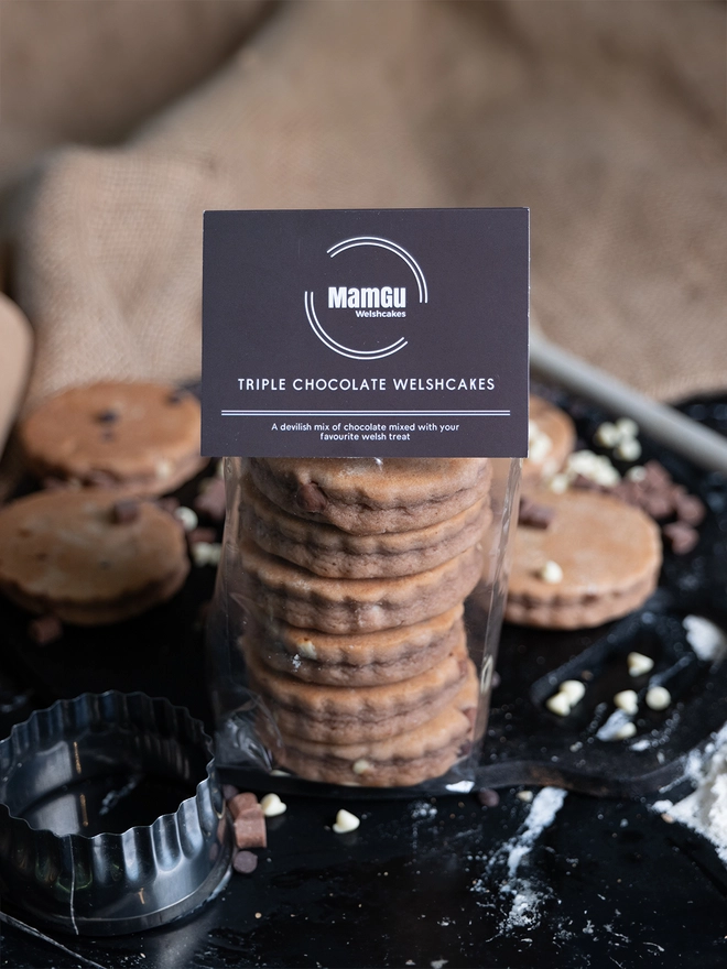 Packaged Triple Chocolate Welsh Cakes with a brown MamGu Welshcakes label with a hessian backdrop and loose welsh cakes on a griddle covered in chocolate chips