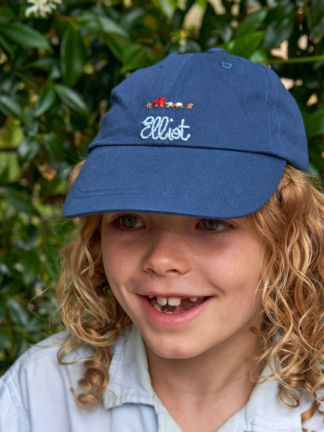 Personalised hand embroidered children's cap. A blue cap with a boys name and a train embroidered on the front