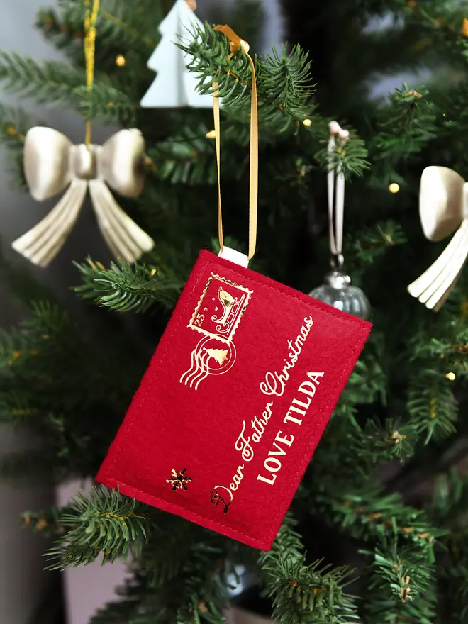 A red felt personalised Christmas envelope that read "Dear Father Christmas, Love Tilda," is hanging on a decorated Christmas tree with golden ribbon.