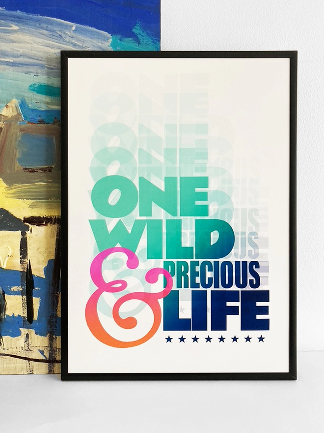 Framed multicoloured typographic print of the last line From Mary Oliver’s poem, The Summer Day - “One Wild And Precious Life”  The print rests against a blue and yellow abstract painting.