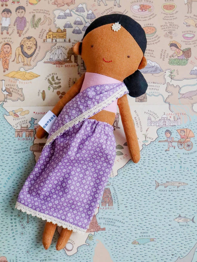 Handmade fabric doll with medium brown skin tone, wearing a lilac patterned sari with lace trim and a pink top. The doll has black hair styled in a low side bun with a small white flower accessory on her forehead, simple stitched facial features. 