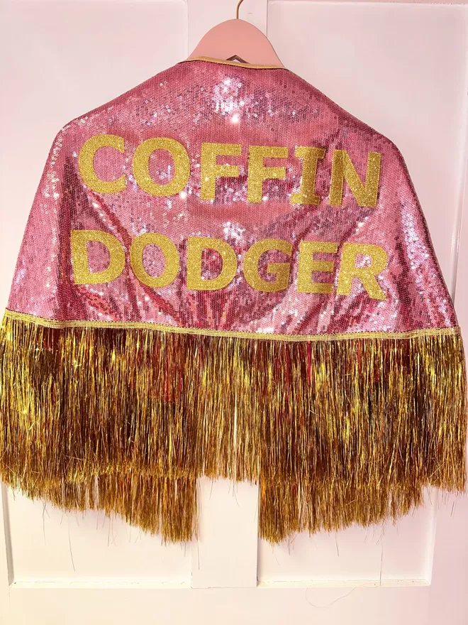 a midi cape with pale pink sequins, gold text reading 'COFFIN DODGER' and gold tinsel
