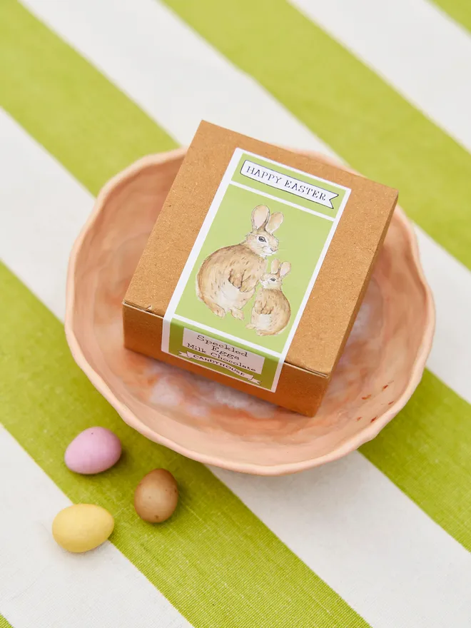 Box of Easter speckled eggs green label places in a pink bowl on a green and whited stripped tablecloth 