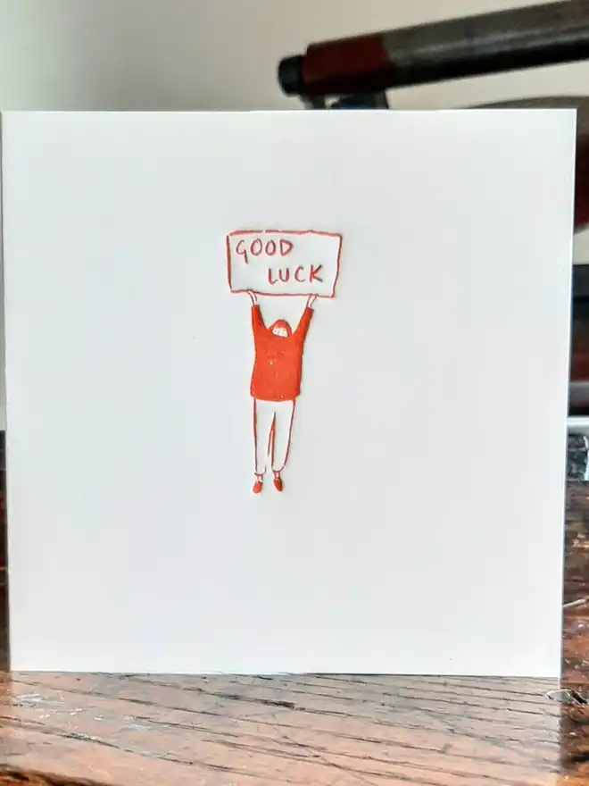 'Good Luck' Letterpress Handprinted Greetings Card, a white good luck card with an orange design on a wooden surface. 