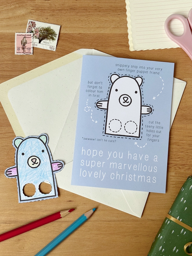 A colour in Christmas card with a polar bear finger puppet design lays on a white envelope on a wooden desk.