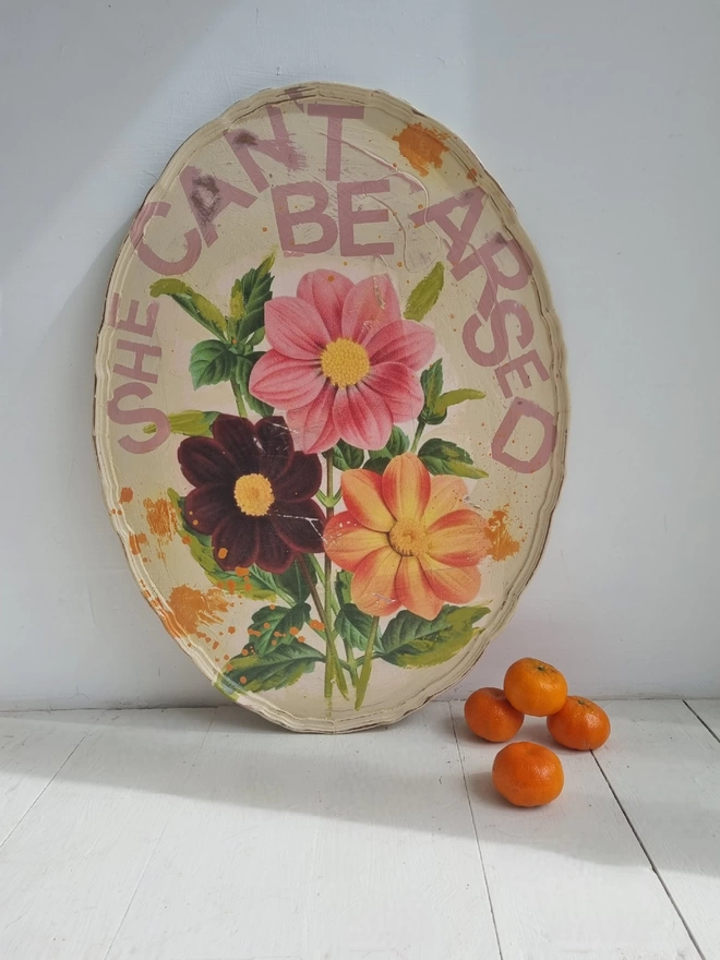 A pie-crust coffee table top upcycled into wall art. Decoupage image of flowers in pinks and oranges taken from a Victorian seed packet. The wording reads in pink, ‘she can’t be arsed’ and the overall style is cream shabby chic. 