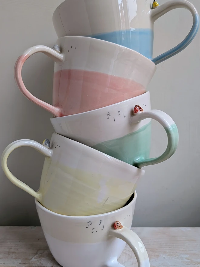 a stack of bright pastel bird cups in blue, pink, green, yellow and white each with a tiny robin or blue tit bird on the handle and musical notes and bird prints