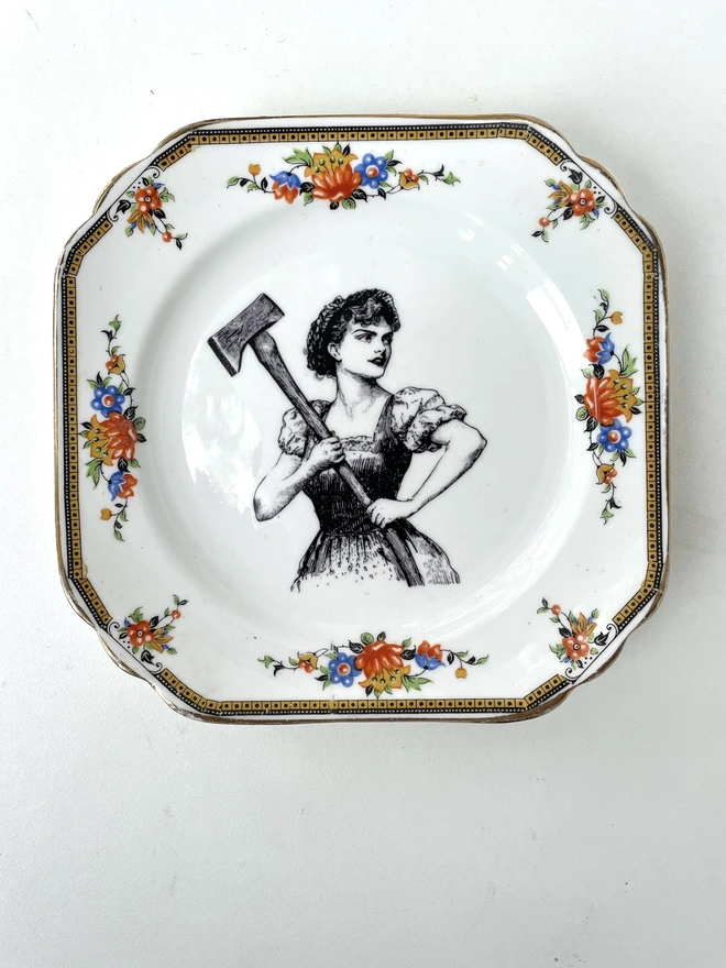 A vintage plate with a patterned border , in the centre of the plate is a black and white vintage illustration of a beautiful lady in a floral dress wielding an axe 