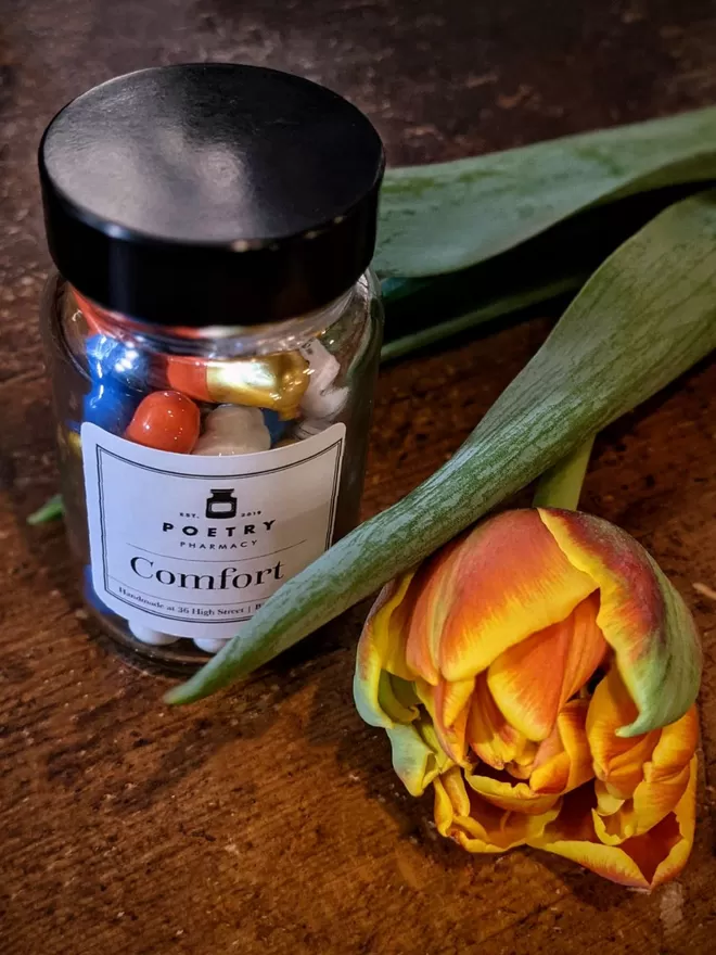A glass bottle of colourful comfort poetry pills with an orange tulip