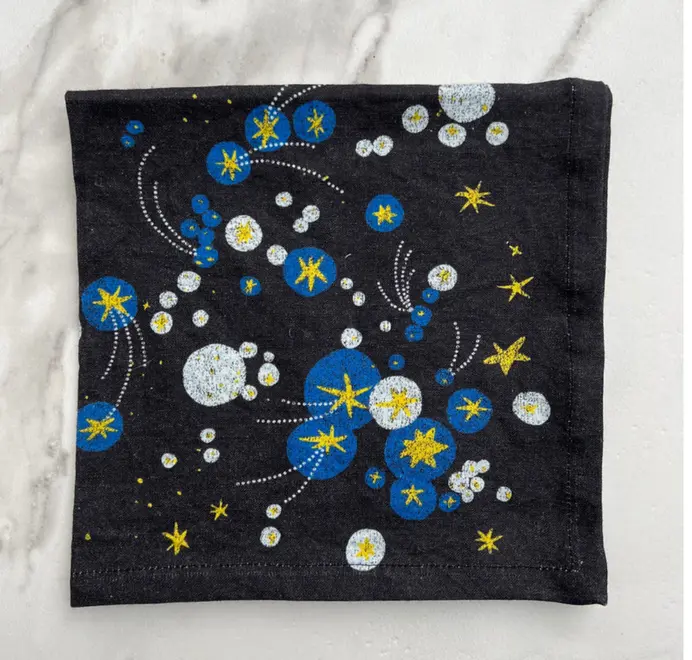 Solaris Napkins In White Or Blue. Blue cloth napkin with blue and yellow starburst and orbital patterns. 