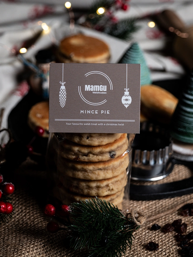 6 Mince Pie Welsh Cakes packed with a a brown christmas bag topper, surrounded by a busy christmas themed kitchen work top, with a bakestone, welsh cake cutter, welsh cakes, and christmas decorations