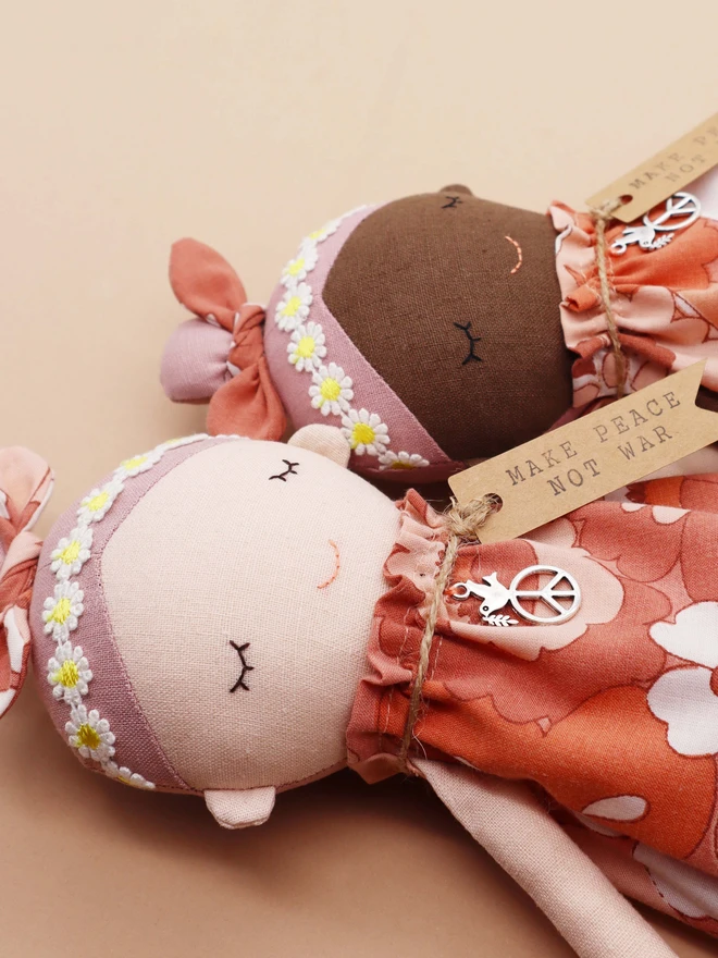 Two handmade fabric dolls called Peace Wishing Flower Child Dolls, each wearing peach and white retro floral dresses, daisy headbands, and bow topknots. They have serene embroidered faces, peace sign charms around their necks, and tags reading “Make Peace Not War.” One doll has a light complexion and the other has a darker complexion, both held side by side against a plain background.