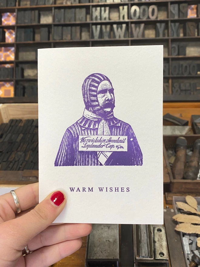 Hand holds up white greeting card with a printed purple illustration and text.