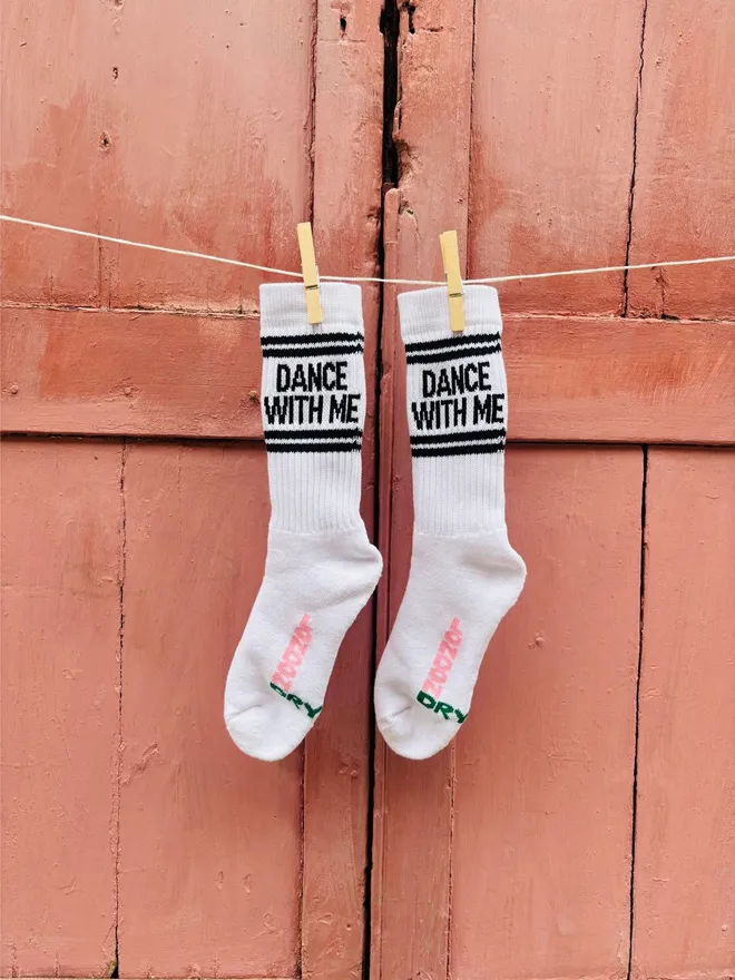 Socks with DANCE WITH ME text shown hung by clothes pegs on washing line in front of distressed pink shutters