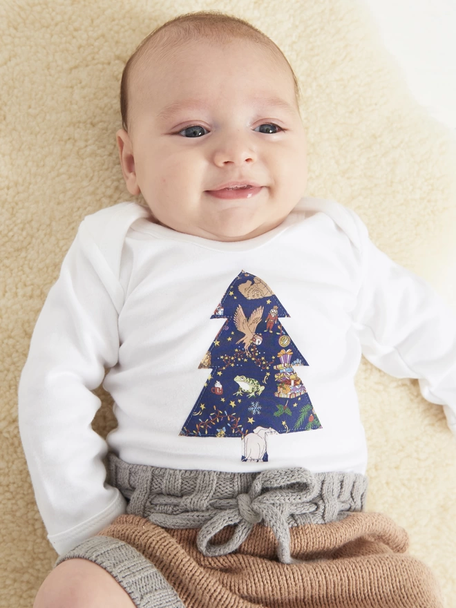 A baby wearing a white bodysuit with a Liberty Christmas festive print Christmas Tree on it