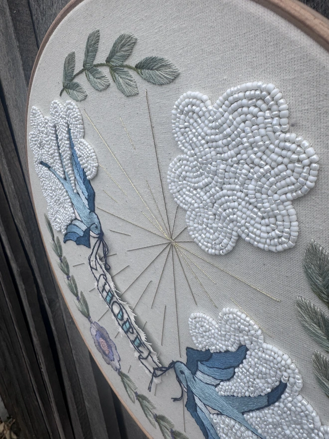 An angled view of an embroidery hoop containing stitched swallows, beaded clouds and sun rays