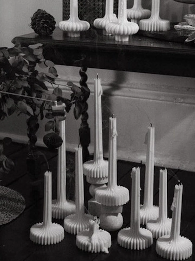 Pair Of White Flair Candles (Set Of 2). A black and white image of tall and short fluted candles clustered on the floor and arranged on a table above, with foliage and a pine cone. The timeless sculptural shapes suit both classic and contemporary interiors.