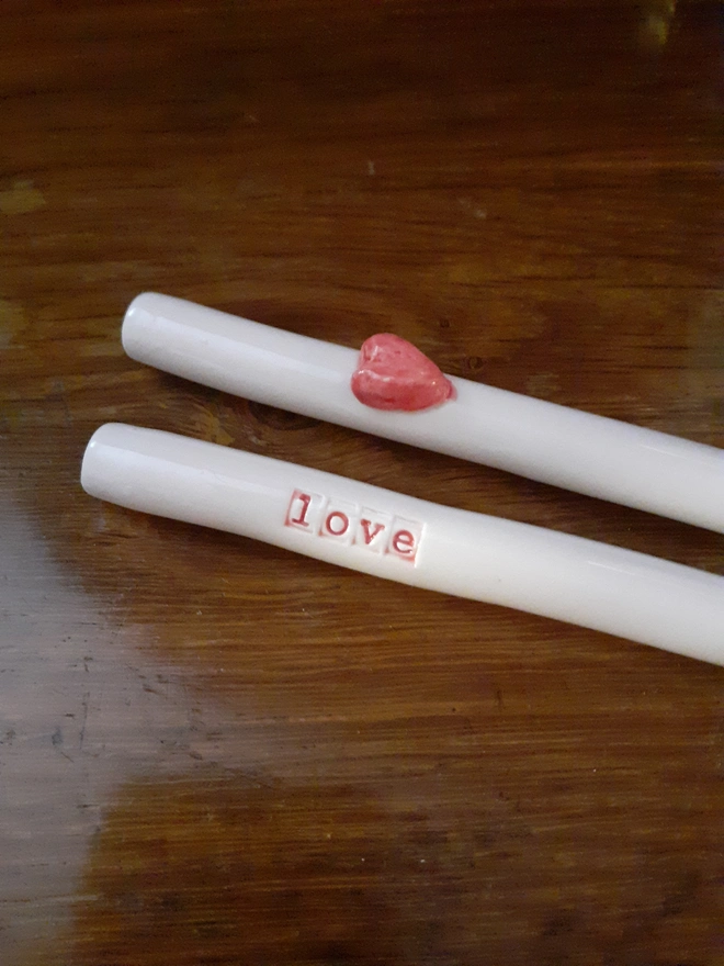 A pair of white ceramic drinking straws, one with the word 'love' inread and the other with a red 3d heart motif