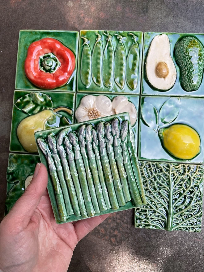 Asparagus tile - square, 3D, realistic and glossy - showing small tightly packed spears, viewed from the side, with lush dark green glaze with purple tips. Other fruit and vegetable tiles in the series are on display in the background: lemon, fig, savoy cabbage, red capsicum pepper, pear, mange tout, avocado, garlic.