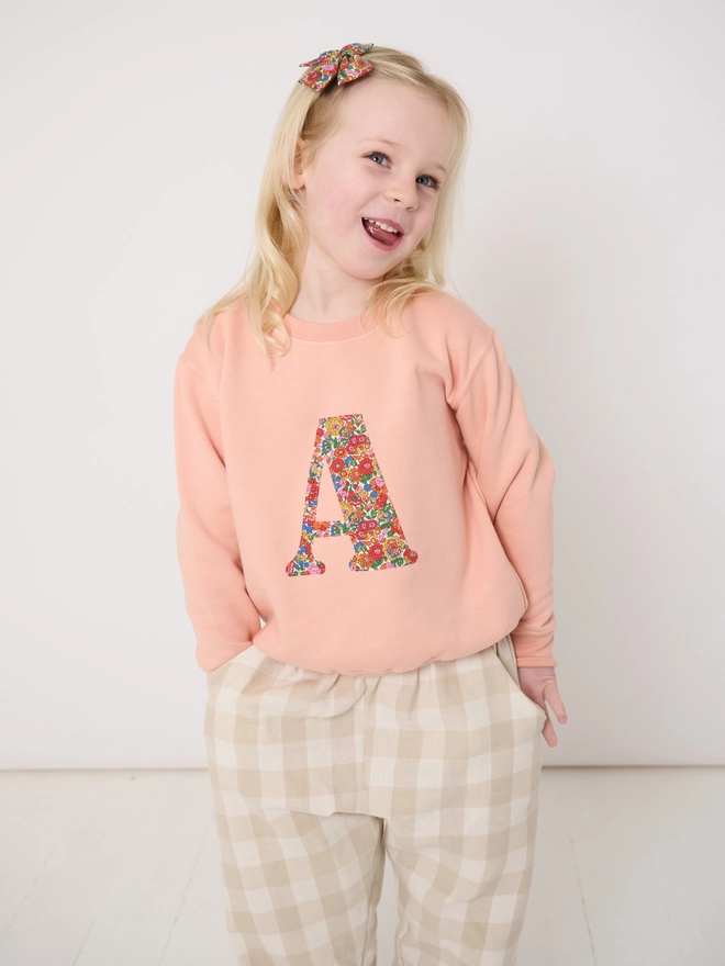 a 5 year old girl wearing a personalised dusty pink sweatshirt with her initial sewn on the front in floral liberty print 