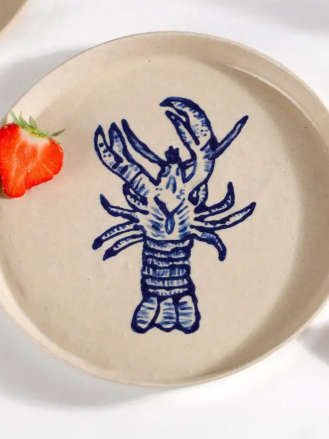 Handmade Ceramic Side Plate With Blue Lobster Design, a side plate with a blue lobster design sitting on a white table surface accompanied by a raspberry. 