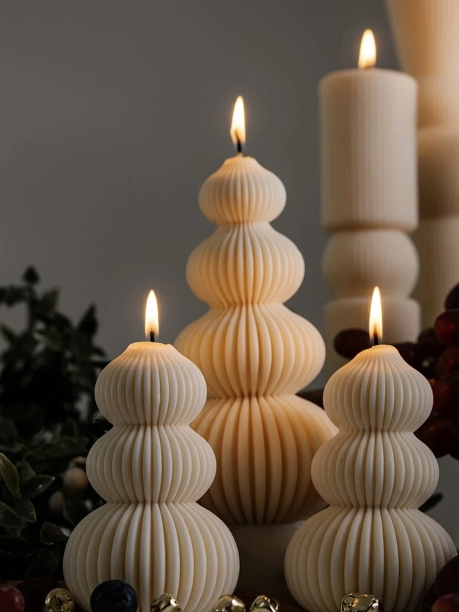 Signature Christmas Tree Sculpture Candle. Close-up of three glowing ribbed candles shaped like stacked baubles in graduating sizes, with gold bells at their base. The fine vertical ridges catch the candlelight beautifully, giving them a modern, luxurious finish.