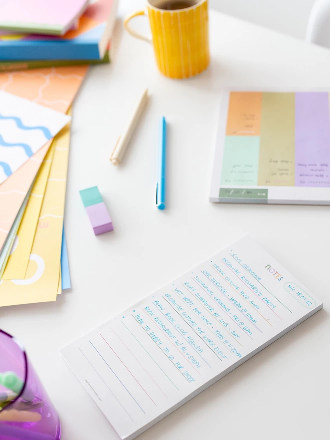 The Raspberry Blossom rainbow coloured lined note pad sits on a desk in a pile of other colourful stationery items from the Happiness collection