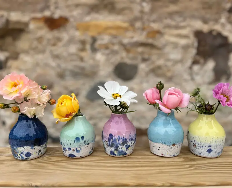 Shoreline Bud Vase, a collection of colourful vases placed on a wooden surface against a brick backdrop. They are accompanied by flowers. 