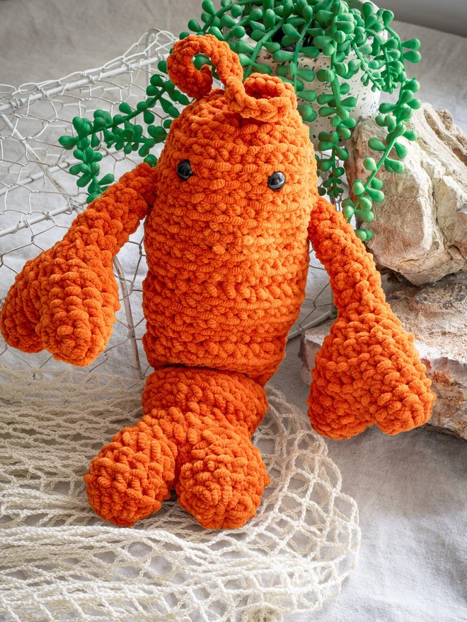 Chunky Chenille Cuddly Crochet Crustacean Lobster Plush, an orange lobster plush sitting accompanied by a bag and some greenery. 