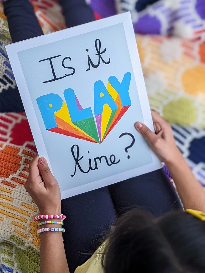 Young girl sitting holding an art print saying 'Is it play time?'