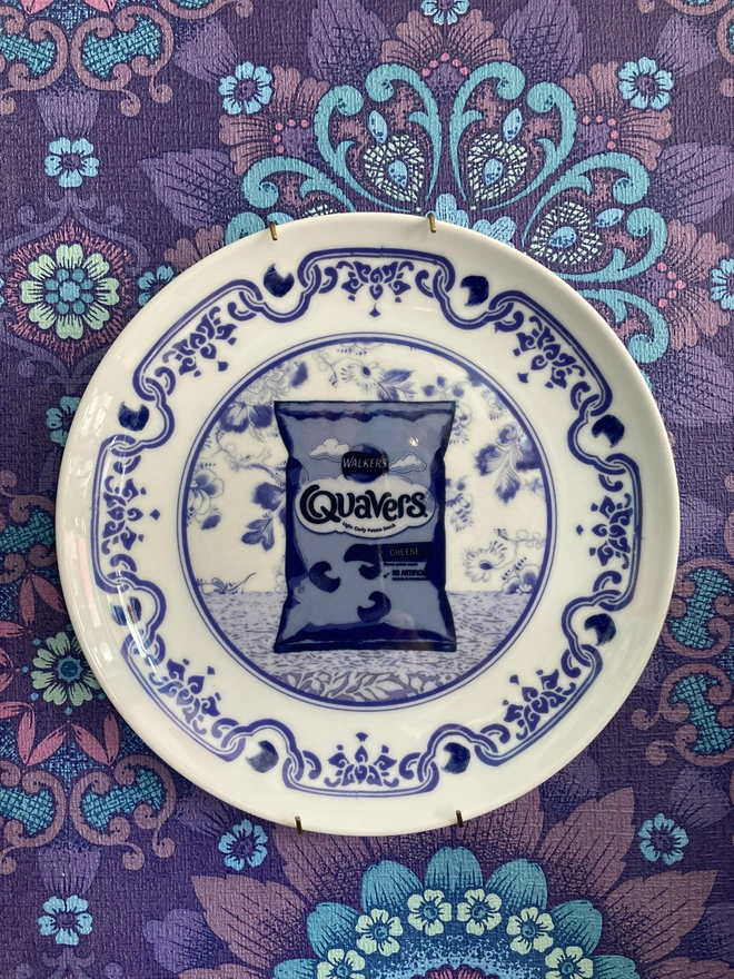 Blue and white Delft style decorative plate featuring a packet of Quavers crisps in the centre.