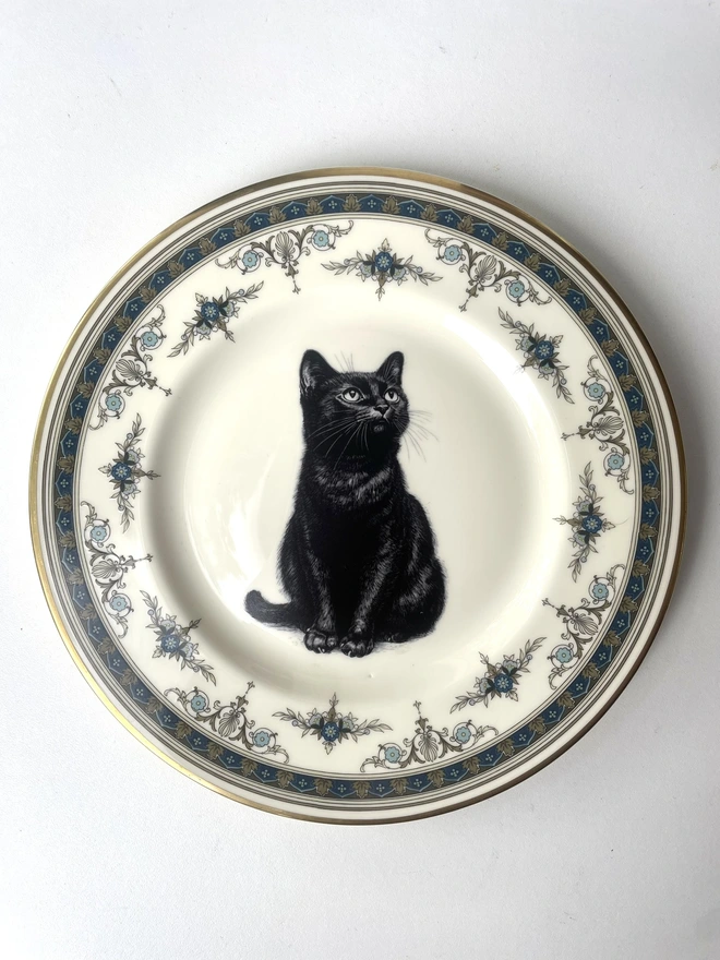 a vintage plate with an ornate border, in the centre of the plate is a vintage style black and white illustration of a black witch's cat