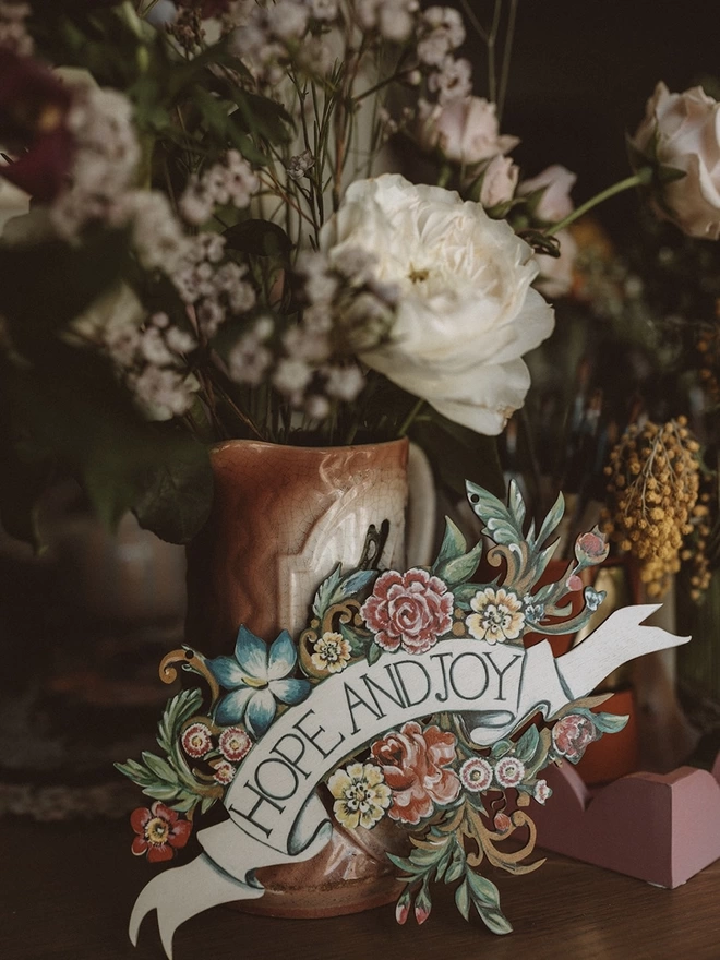 Wooden banner saying 'Hope And Joy' nestled in flowers, displayed with fresh flowers