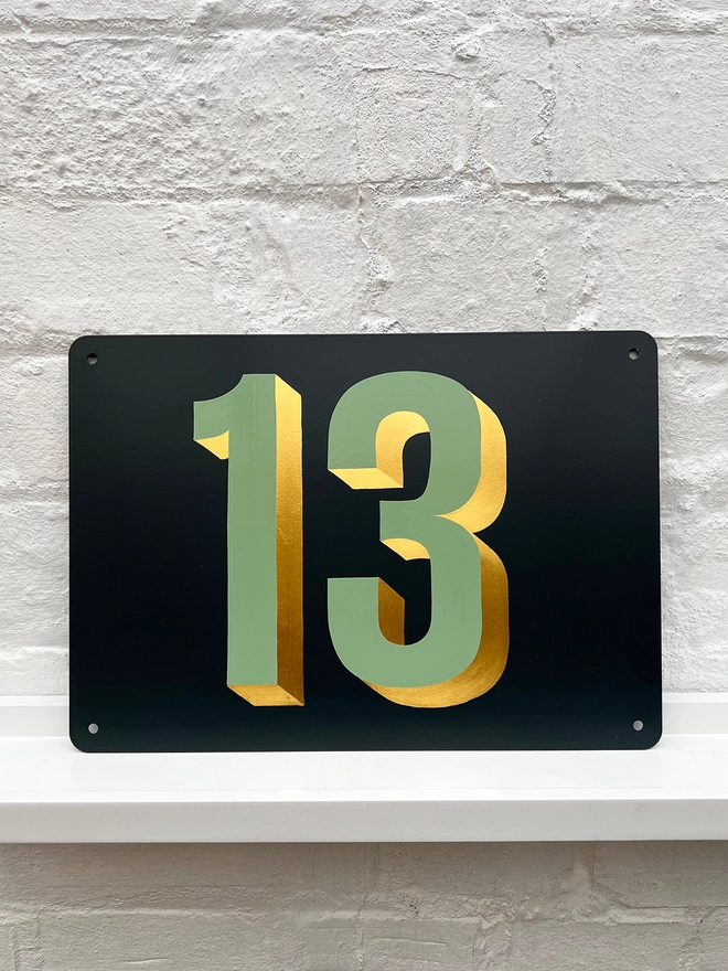 Sage green and gold leaf house number 13, on anthracite grey metal plaque, against a white brick wall.