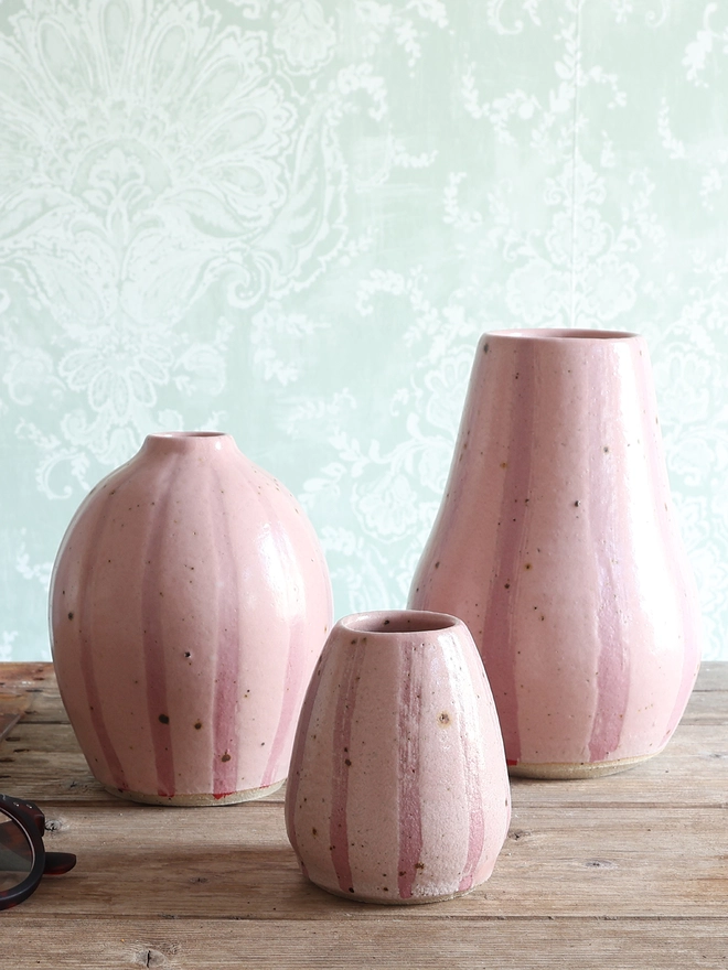 Three Handmade Ceramic Stoneware Vases with a pink stripe design sat on a wooden surface next to a pair of glasses. A medium rounded vase sits of the left, with a large vase next to it and a small bud vase in-between. 