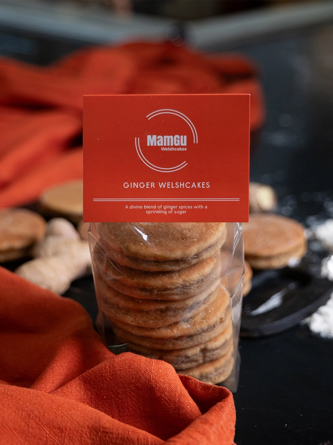 Packaged Ginger Welsh Cakes with an Orange MamGu Welshcakes label, busy worktop background on an orange table cloth