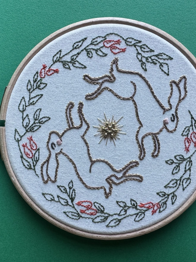 An embroidery hoop containing two hares edged in gold circling a gold bead and contained by leaves and rose hips